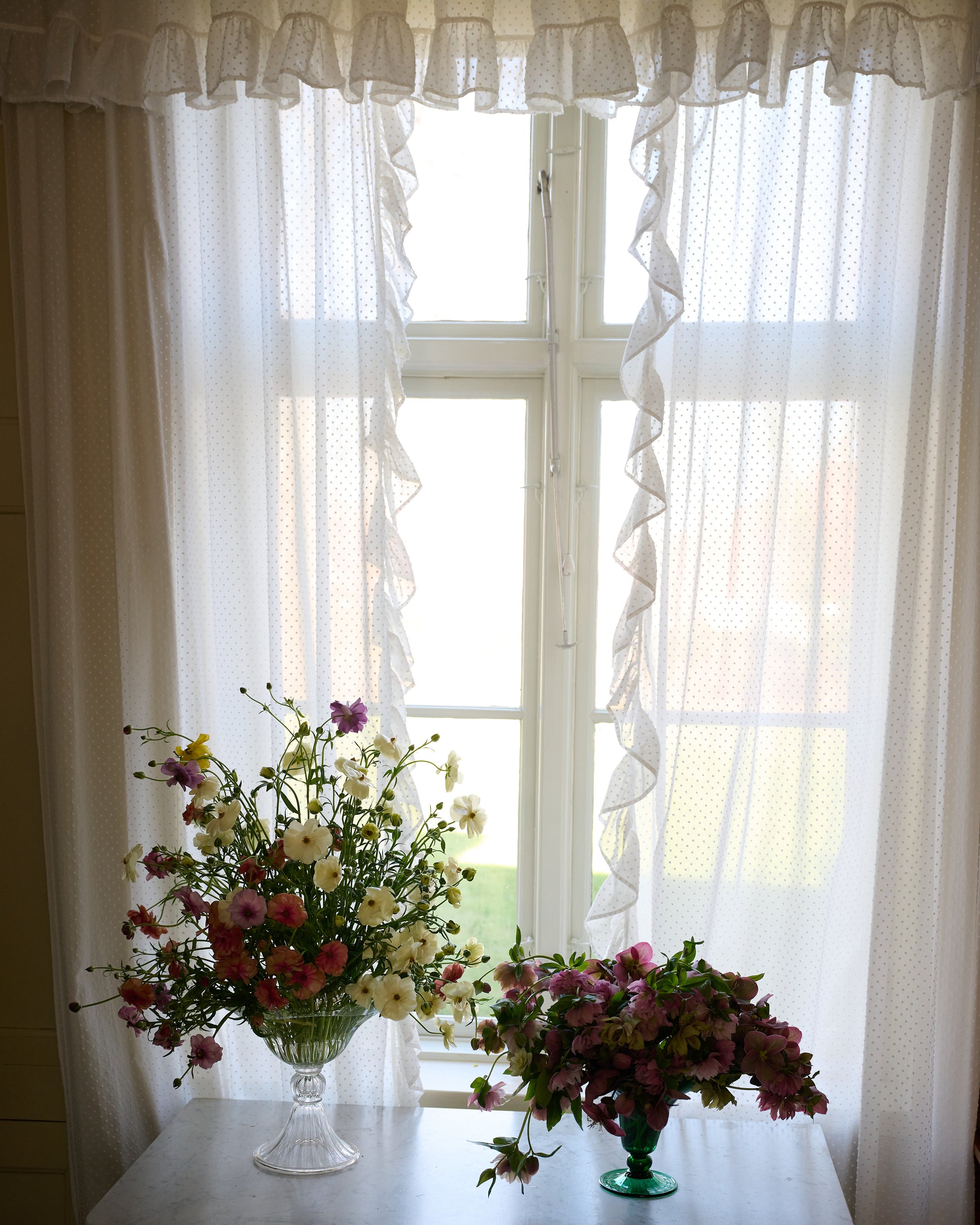 Small and large mouth blown green and clear Murano vases in window with flowers.