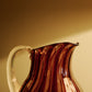 A striped glass pitcher with in amber and pink with a clear handle. 