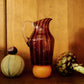 A striped glass pitcher with amber and pink hues and a clear handle, surrounded by vegetables.