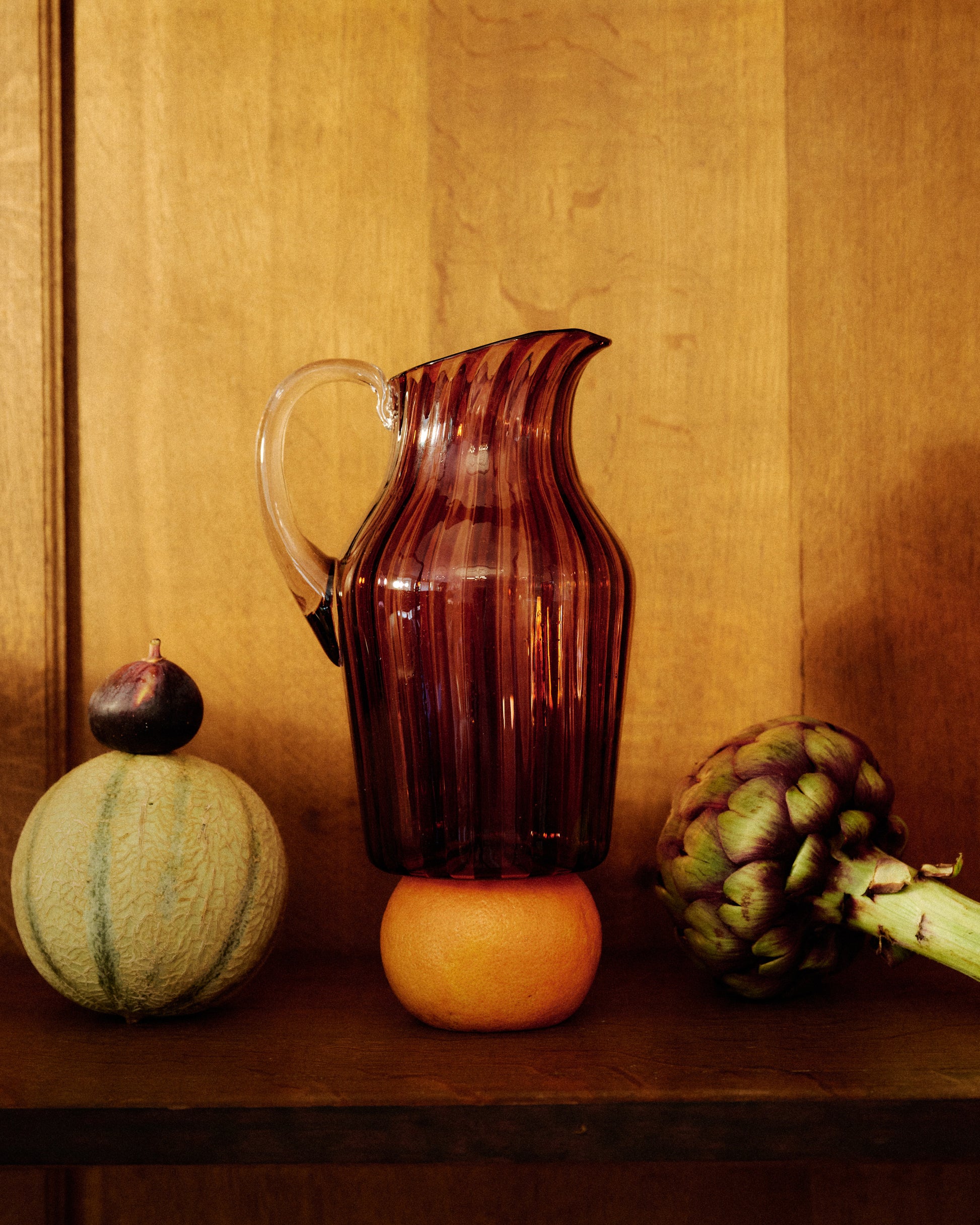 A striped glass pitcher with amber and pink hues and a clear handle, surrounded by vegetables.