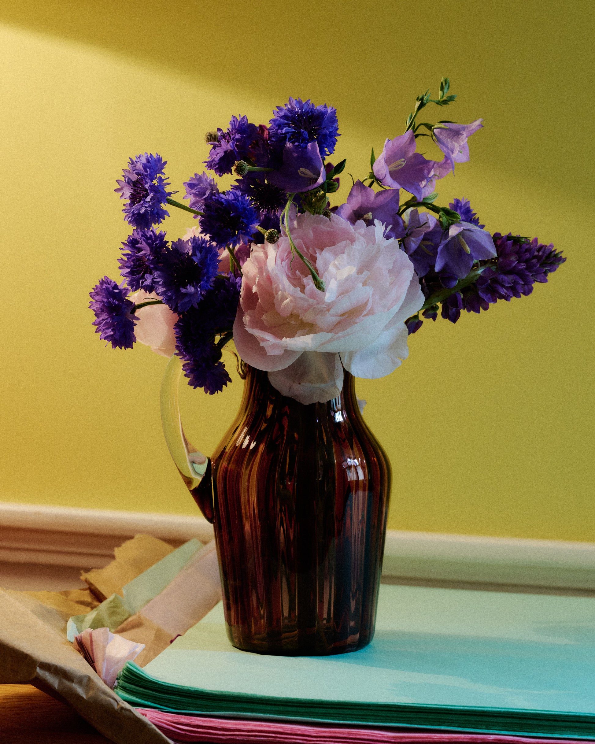 A striped glass pitcher with flowers in amber and pink and a clear handle. 