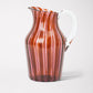 A striped glass pitcher with amber and pink hues and a clear handle.